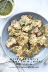 A gray plate of roasted smashed potatoes topped with creamy chimichurri sauce sits next to a bowl of extra sauce and a whisk. Text at the bottom reads Macro-Friendly Creamy Chimichurri Dressing.
