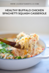 A wooden spoon lifts a serving of Buffalo Chicken Spaghetti Squash Casserole from a white dish, the casserole topped with fresh green herbs for a vibrant finishing touch.
