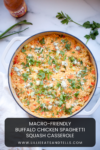 A large white casserole dish filled with Buffalo Chicken Spaghetti Squash Casserole, topped with fresh herbs and crumbled blue cheese, sits on a marble surface beside sprigs of cilantro, parsley, and a bottle of hot sauce.