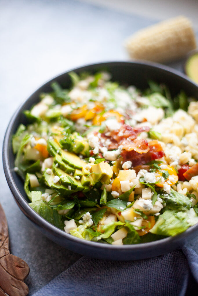 A close-up of a loaded Copycat Factory Chopped Salad in a dark bowl, featuring chopped greens, avocado, diced cheese, crumbled blue cheese, cherry tomatoes, and crispy bacon, all topped with a creamy dressing.
