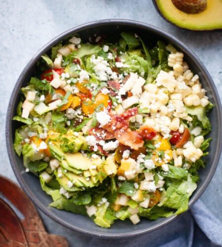 A bowl of salad with lettuce, chopped tomatoes, corn, diced cheese, avocado slices, bacon bits, and crumbled cheese—just like a Copycat Factory Chopped Salad. Half an avocado is nearby.