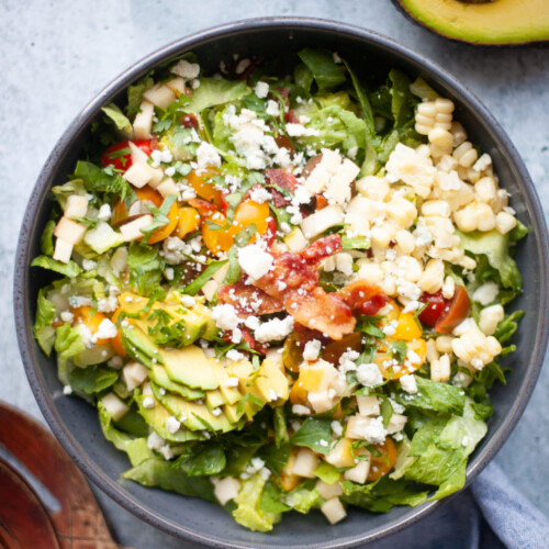 A bowl of salad with lettuce, chopped tomatoes, corn, diced cheese, avocado slices, bacon bits, and crumbled cheese—just like a Copycat Factory Chopped Salad. Half an avocado is nearby.