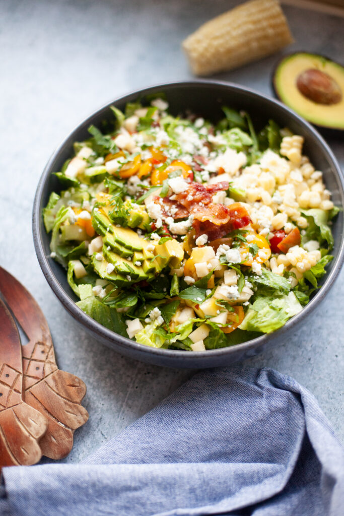 A bowl of Copycat Factory Chopped Salad features crisp lettuce, creamy avocado, cheese, bacon, and fresh vegetables, all topped with crumbled cheese. Wooden salad tongs, a blue napkin, corn, and half an avocado are nearby.