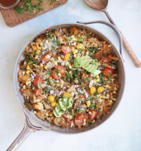 A skillet filled with a colorful BBQ Chicken and Butternut Macro-Friendly Skillet, topped with sliced avocado, cherry tomatoes, corn, onion, melted cheese, and chopped cilantro, with wooden utensils nearby.