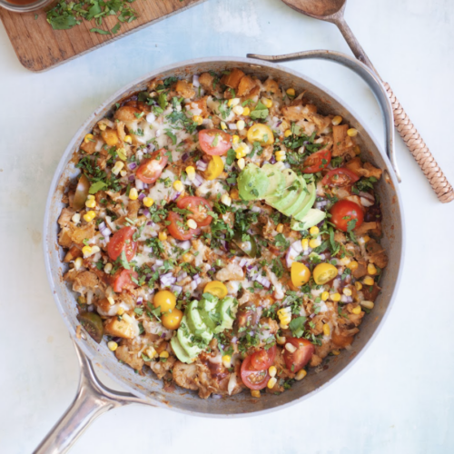 A skillet filled with a colorful BBQ Chicken and Butternut Macro-Friendly Skillet, topped with sliced avocado, cherry tomatoes, corn, onion, melted cheese, and chopped cilantro, with wooden utensils nearby.