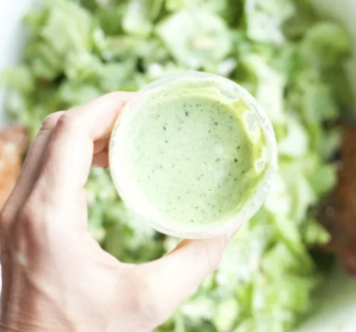 A hand holds a small container of creamy green Jalapeño-Avocado Dressing above a bowl of chopped green lettuce.