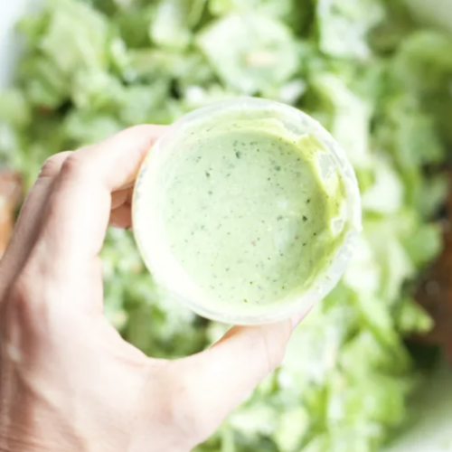 A hand holds a small container of creamy green Jalapeño-Avocado Dressing above a bowl of chopped green lettuce.