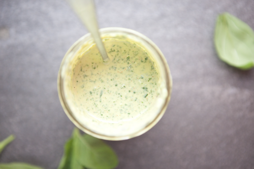 A glass jar filled with creamy green herb sauce, viewed from above, with a spoon inside. This delicious Creamy Chimichurri is garnished with vibrant basil leaves scattered on the gray surface around the jar.