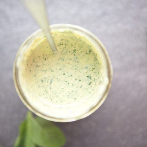 A glass jar filled with creamy green herb sauce, viewed from above, with a spoon inside. This delicious Creamy Chimichurri is garnished with vibrant basil leaves scattered on the gray surface around the jar.