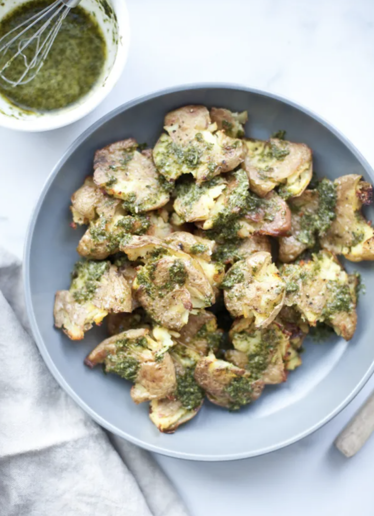 A blue plate filled with smashed roasted potatoes topped with green pesto sauce. A bowl of extra creamy chimichurri and a whisk are nearby on a white surface with a beige napkin.