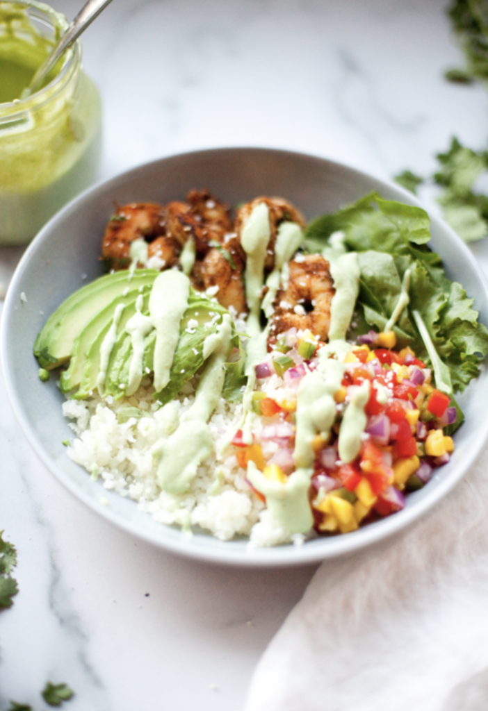 A bowl with grilled shrimp, avocado slices, greens, cauliflower rice, colorful salsa, and green Jalapeño-Avocado Dressing drizzled on top sits on a white surface with a jar of dressing and scattered herbs nearby.