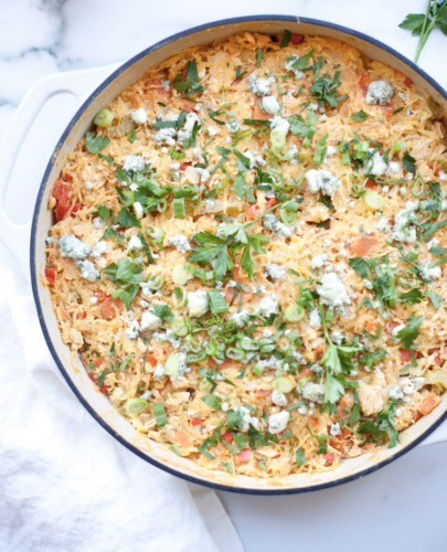 A large white pan filled with Buffalo Chicken Spaghetti Squash Casserole is topped with chopped green onions, parsley, and crumbled blue cheese, with visible bits of chicken and vegetables mixed in. A white napkin sits next to the pan.