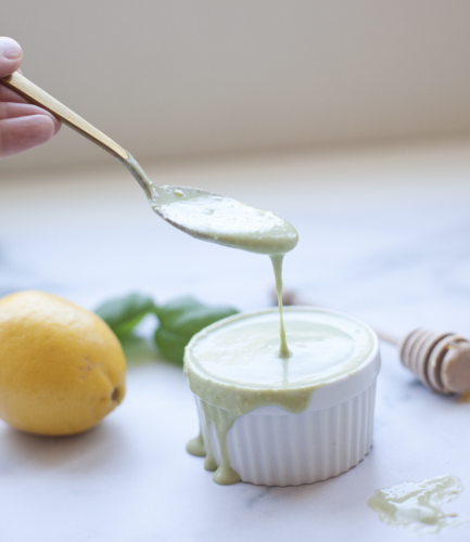 A spoon drizzles Creamy Lemon-Basil Dressing into a white ramekin, with the sauce overflowing. A lemon, basil leaves, and a honey dipper are blurred in the background on a marble surface.