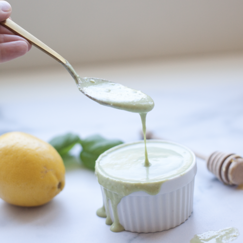 A spoon drizzles Creamy Lemon-Basil Dressing into a white ramekin, with the sauce overflowing. A lemon, basil leaves, and a honey dipper are blurred in the background on a marble surface.