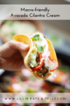A hand holds a taco filled with vegetables, topped with zesty Avocado-Cilantro Cream, onion, tomato, and greens. Text above reads Macro-friendly Avocado Cilantro Cream and website link is shown below.