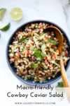 A bowl of colorful Macro-friendly Cowboy Caviar Salad with chopped vegetables and beans, garnished with fresh cilantro and served with a wooden spoon. Sliced limes sit beside the bowl on a white surface. Text reads: Macro-friendly Cowboy Caviar Salad.