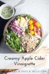 A bowl filled with arugula, quinoa, sliced apples, tomatoes, onion, chicken, cheese, and nuts is paired perfectly with a spoon and jar of Creamy Apple Cider Vinaigrette on the side. Text: Creamy Apple Cider Vinaigrette, www.lillieeatsandtells.com.