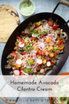 Oval pan with sliced beef, cherry tomatoes, peppers, onions, corn, and cheese, garnished with cilantro. A small bowl of fresh Avocado-Cilantro Cream and a round cutting board are next to the pan. Text reads: Homemade Avocado Cilantro Cream.
