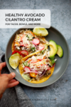 A hand holds a gray bowl with two tacos topped with slaw, radishes, avocado, and lime wedges. Text reads: Healthy Avocado-Cilantro Cream for tacos, bowls, and more. Discover this delicious Avocado-Cilantro Cream recipe at lillieeatsandtells.com.