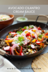 A skillet filled with sliced beef, colorful bell peppers, radishes, corn, onions, and a dollop of creamy Avocado-Cilantro Cream sauce sits alongside warm tortillas and a fresh salad bowl. Text reads Avocado Cilantro Cream Sauce.