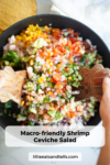 A bowl of colorful Shrimp Ceviche Salad is mixed with wooden salad tongs. The dish includes diced tomatoes, avocado, cucumber, red onion, and herbs. Text on the image reads, Macro-friendly Shrimp Ceviche Salad.