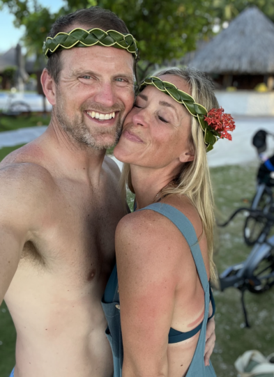 A smiling couple wearing leaf crowns poses for a selfie outdoors. The woman has a red flower in her hair; both appear sunburned and happy, with tropical scenery and a thatched building in the background—capturing the joy of How We Planned the Trip to Moorea and Bora Bora.