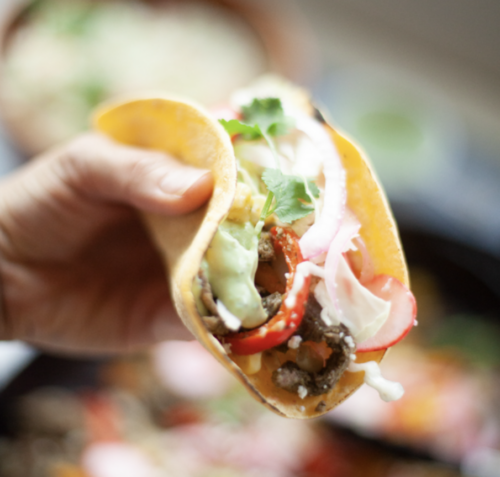 A hand holds a soft taco filled with beef, sliced red peppers, onions, lettuce, and topped with cilantro and a generous drizzle of Avocado-Cilantro Cream.