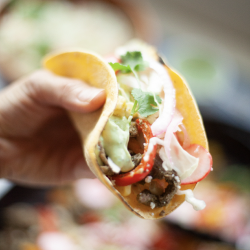 A hand holds a soft taco filled with beef, sliced red peppers, onions, lettuce, and topped with cilantro and a generous drizzle of Avocado-Cilantro Cream.