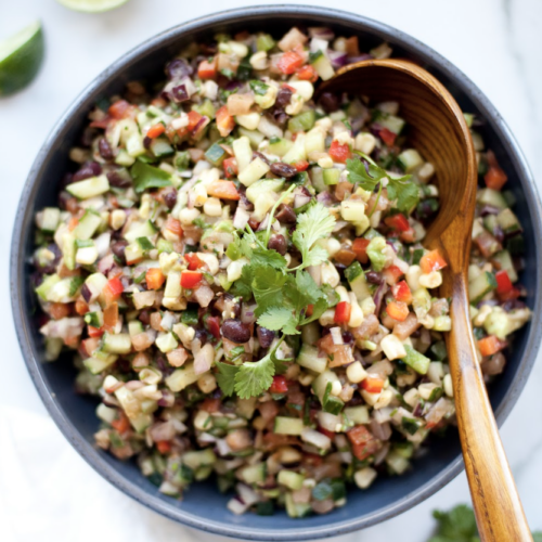 A bowl of colorful chopped salad with black beans, corn, cucumber, peppers, and cilantro, with a wooden spoon resting inside—this vibrant dish is a delicious example of Macro-friendly Cowboy Caviar Salad.