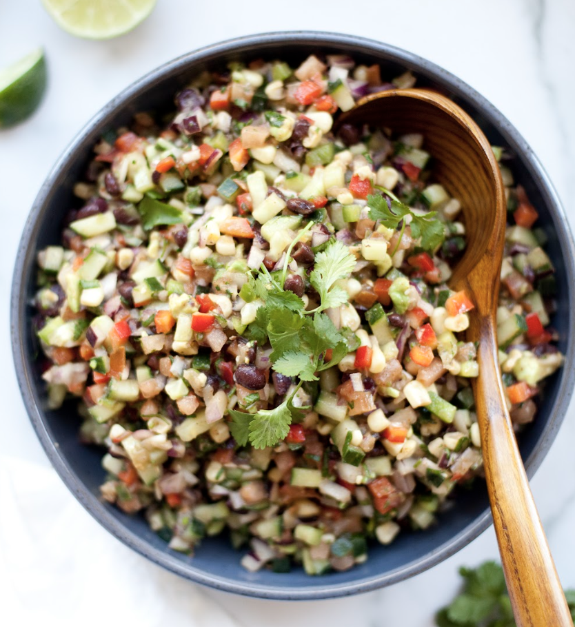 A bowl of colorful chopped salad with black beans, corn, cucumber, peppers, and cilantro, with a wooden spoon resting inside—this vibrant dish is a delicious example of Macro-friendly Cowboy Caviar Salad.