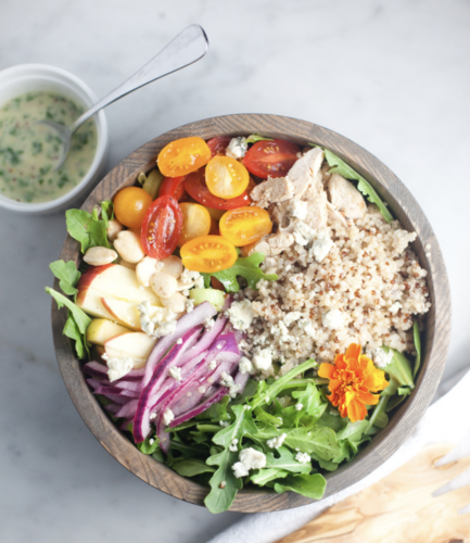 A wooden bowl filled with arugula, cherry tomatoes, quinoa, shredded chicken, apple slices, red onion, nuts, blue cheese, and an orange flower is accompanied by a cup of creamy apple cider vinaigrette and a spoon nearby.