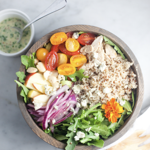 A wooden bowl filled with arugula, cherry tomatoes, quinoa, shredded chicken, apple slices, red onion, nuts, blue cheese, and an orange flower is accompanied by a cup of creamy apple cider vinaigrette and a spoon nearby.