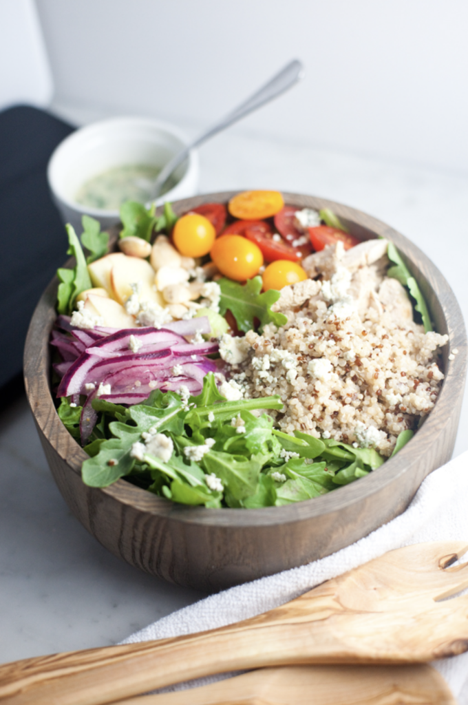 A wooden bowl filled with arugula, quinoa, cherry tomatoes, red onions, cashews, grilled chicken, and cheese crumbles is beautifully presented. Nearby, a small bowl of creamy apple cider vinaigrette and wooden salad tongs complete the inviting scene.