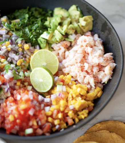 A bowl with chopped avocado, cilantro, red onion, mango, tomato, cucumber, and crab brings to mind the fresh flavors of a Shrimp Ceviche Salad. Two lime slices add a zesty touch while tortilla chips are served next to the bowl for the perfect crunch.