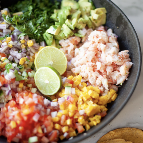 A bowl with chopped avocado, cilantro, red onion, mango, tomato, cucumber, and crab brings to mind the fresh flavors of a Shrimp Ceviche Salad. Two lime slices add a zesty touch while tortilla chips are served next to the bowl for the perfect crunch.