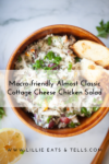 A wooden bowl of almost classic cottage cheese chicken salad is garnished with green onions, olives, and bread slices. A lemon slice and herbs are nearby. Text overlay reads: Macro-friendly Almost Classic Cottage Cheese Chicken Salad.