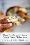 A hand holds a piece of bread topped with almost classic cottage cheese chicken salad, with a wooden bowl of salad and a spoon in the blurred background. Text reads: Macro-friendly Almost Classic Cottage Cheese Chicken Salad.