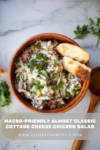 A wooden bowl of almost classic cottage cheese chicken salad with olives and herbs, topped with green onions, served with toasted bread slices. Text reads: Macro-friendly almost classic cottage cheese chicken salad.