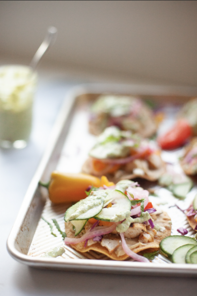 A baking tray with tostadas topped with sliced cucumber, onions, and herbs, served alongside a yellow pepper and a glass jar of creamy Cucumber Dill Sauce in the background.