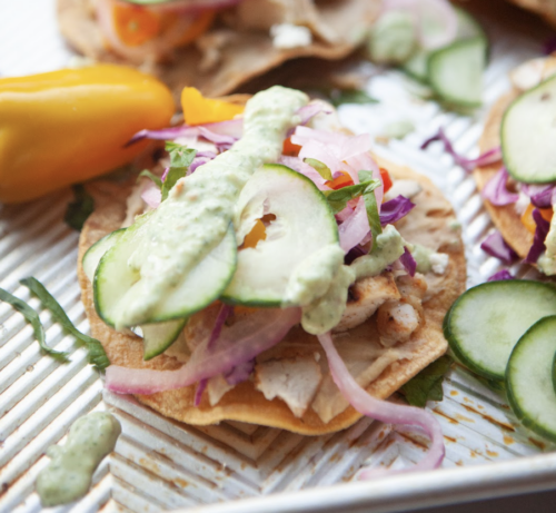 A close-up of tostadas topped with sliced cucumber, red onion, shredded greens, chicken, and a drizzle of creamy Cucumber Dill Sauce, with a yellow pepper on the side.
