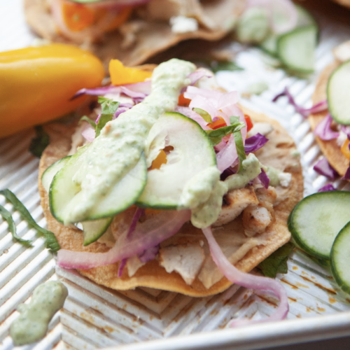 A close-up of tostadas topped with sliced cucumber, red onion, shredded greens, chicken, and a drizzle of creamy Cucumber Dill Sauce, with a yellow pepper on the side.