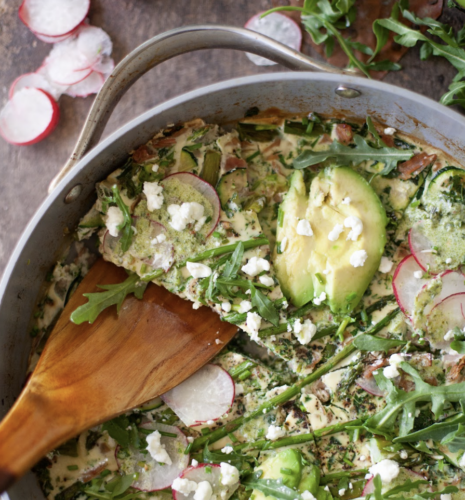 A spring frittata with avocado, radish slices, arugula, asparagus, and crumbled cheese is being served from the skillet with a wooden spatula. Fresh radishes and greens are visible nearby, highlighting the vibrant flavors of the season.