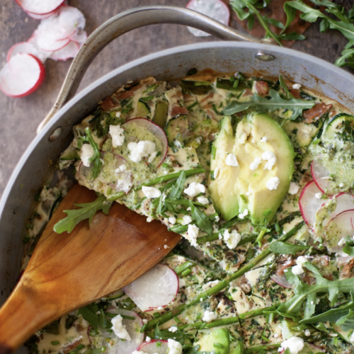 A spring frittata with avocado, radish slices, arugula, asparagus, and crumbled cheese is being served from the skillet with a wooden spatula. Fresh radishes and greens are visible nearby, highlighting the vibrant flavors of the season.