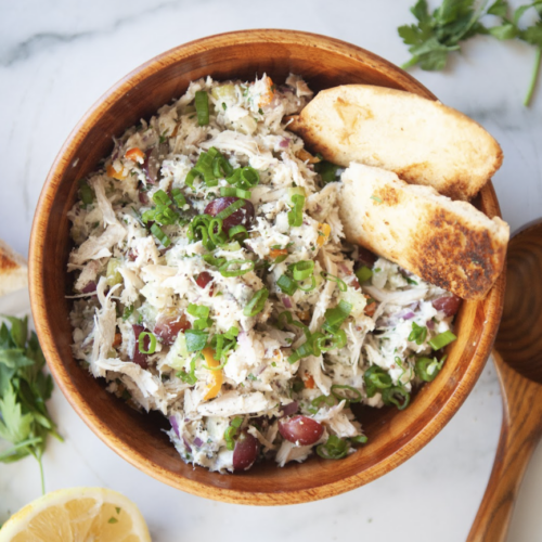 A wooden bowl filled with almost classic cottage cheese chicken salad, garnished with green onions and served with two pieces of toasted bread. Parsley, a lemon slice, and a wooden spoon are nearby on a marble surface.