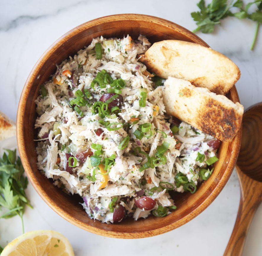 A wooden bowl filled with almost classic cottage cheese chicken salad, garnished with green onions and served with two pieces of toasted bread. Parsley, a lemon slice, and a wooden spoon are nearby on a marble surface.