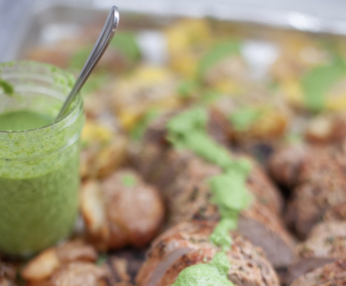 A jar of Jalapeño Herb Sauce with a spoon is placed next to roasted potatoes on a tray, with the vibrant sauce drizzled over the potatoes. The image is slightly out of focus in the background.