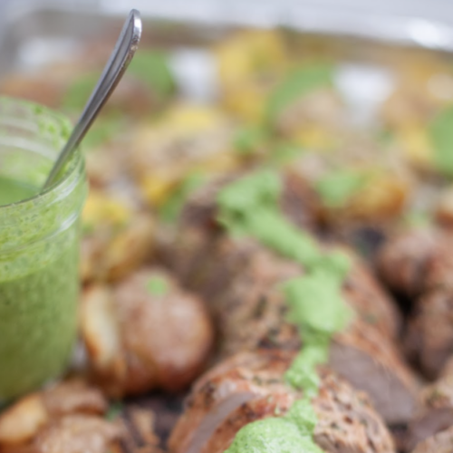A jar of Jalapeño Herb Sauce with a spoon is placed next to roasted potatoes on a tray, with the vibrant sauce drizzled over the potatoes. The image is slightly out of focus in the background.