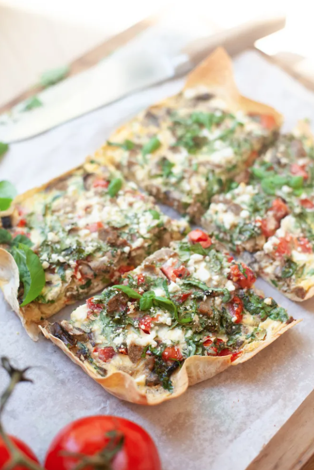 A rectangular veggie pizza with a thin crust, topped with greens, tomatoes, cheese, and herbs, is sliced into squares and served on parchment paper. Fresh tomatoes and basil are nearby—making this one of the top macro-friendly Mother’s Day brunch ideas for a delicious and wholesome celebration.