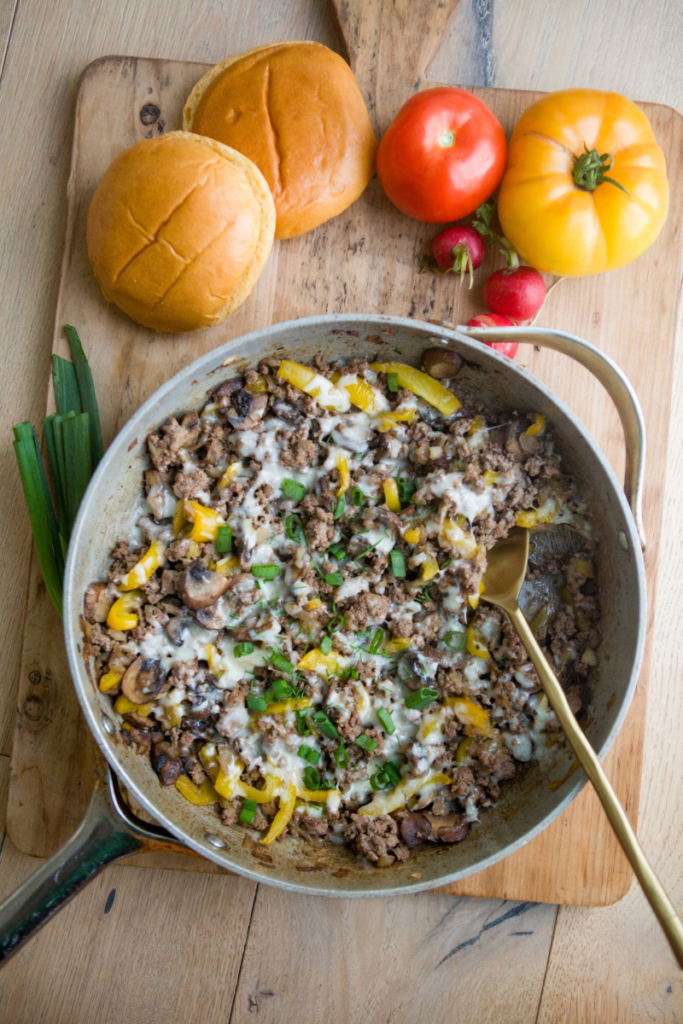 Ground beef philly skillet in large skillet with gold spoon. Buns and tomatoes are off to the side on wooden cutting board and sprig of green onions underneath skillet.