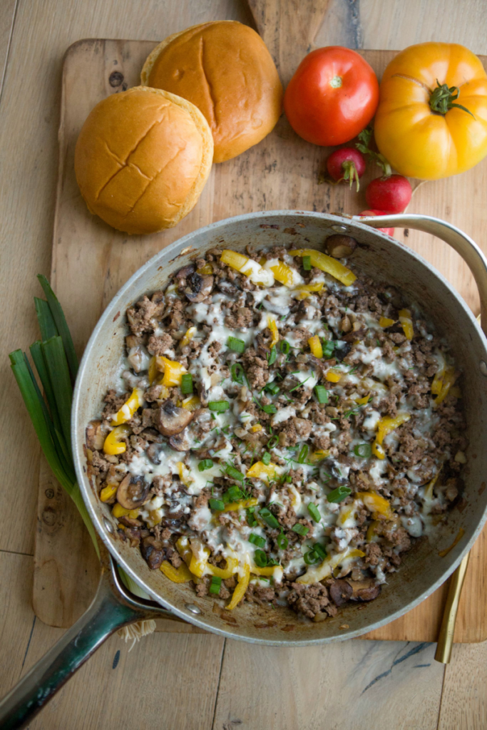 Macro-friendly Ground Beef Philly Skillet in large skillet on wooden cutting board with gold spoon and green onions underneath.